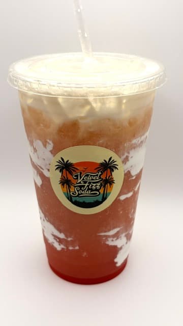 Layered reddish-orange soda with white cream foam in a cup with Velvet Fizz Soda logo.